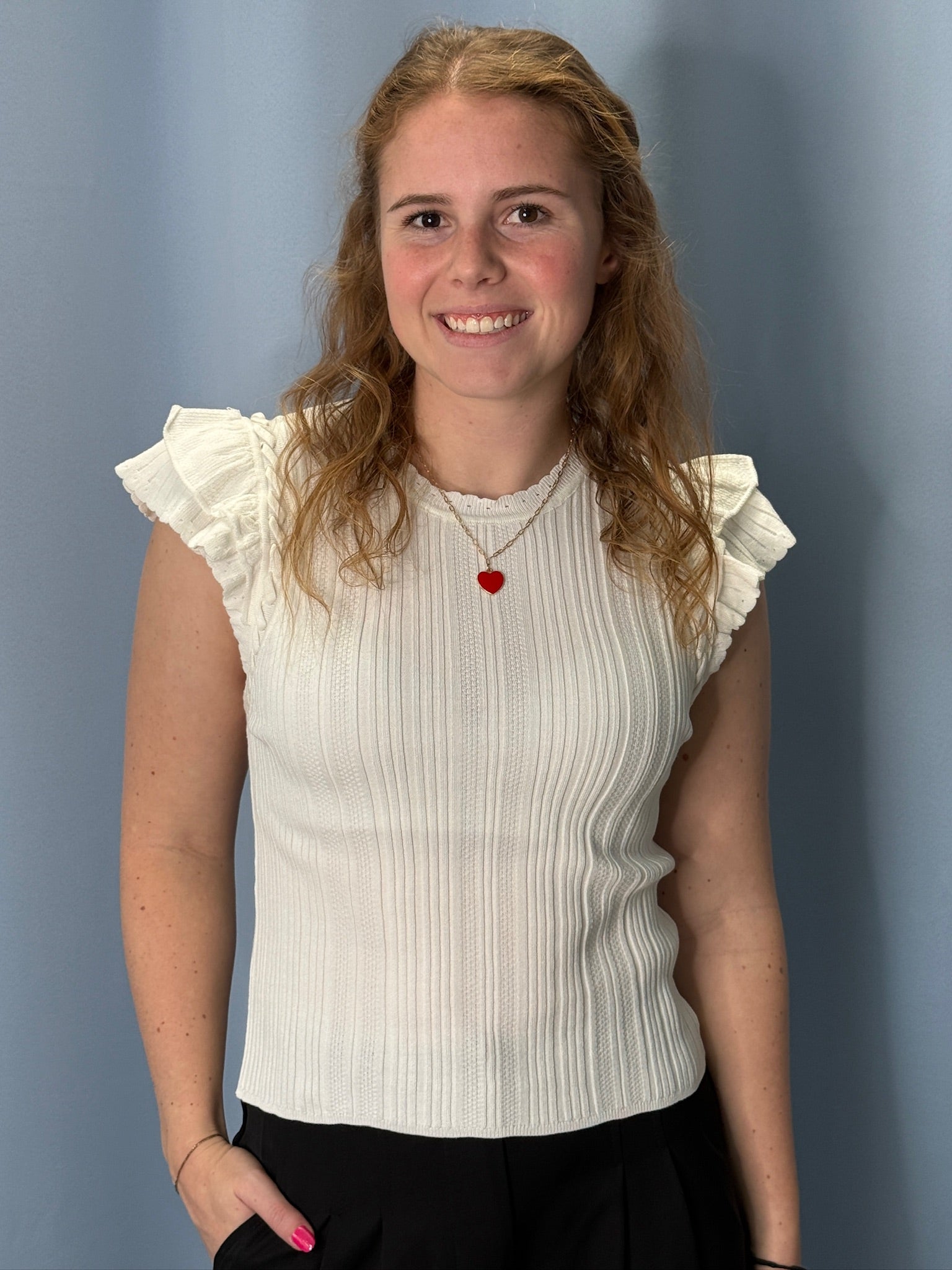 Woman wearing a white ruffled sleeve top with a red heart pendant against a plain background