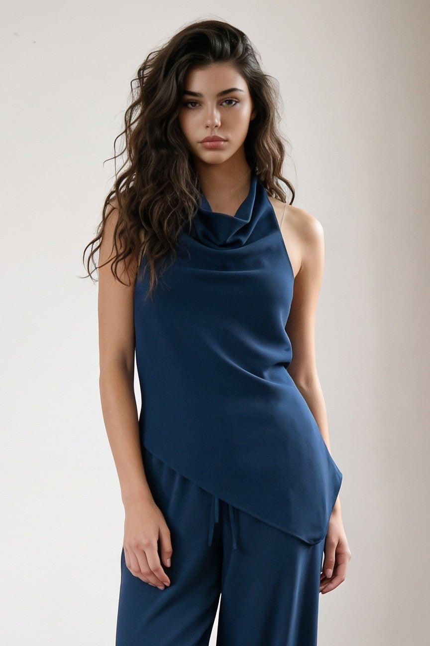 A woman modeling a sleeveless navy blue cowl-neck top with an asymmetrical draped hem, paired with matching navy pants, against a plain light beige studio background.