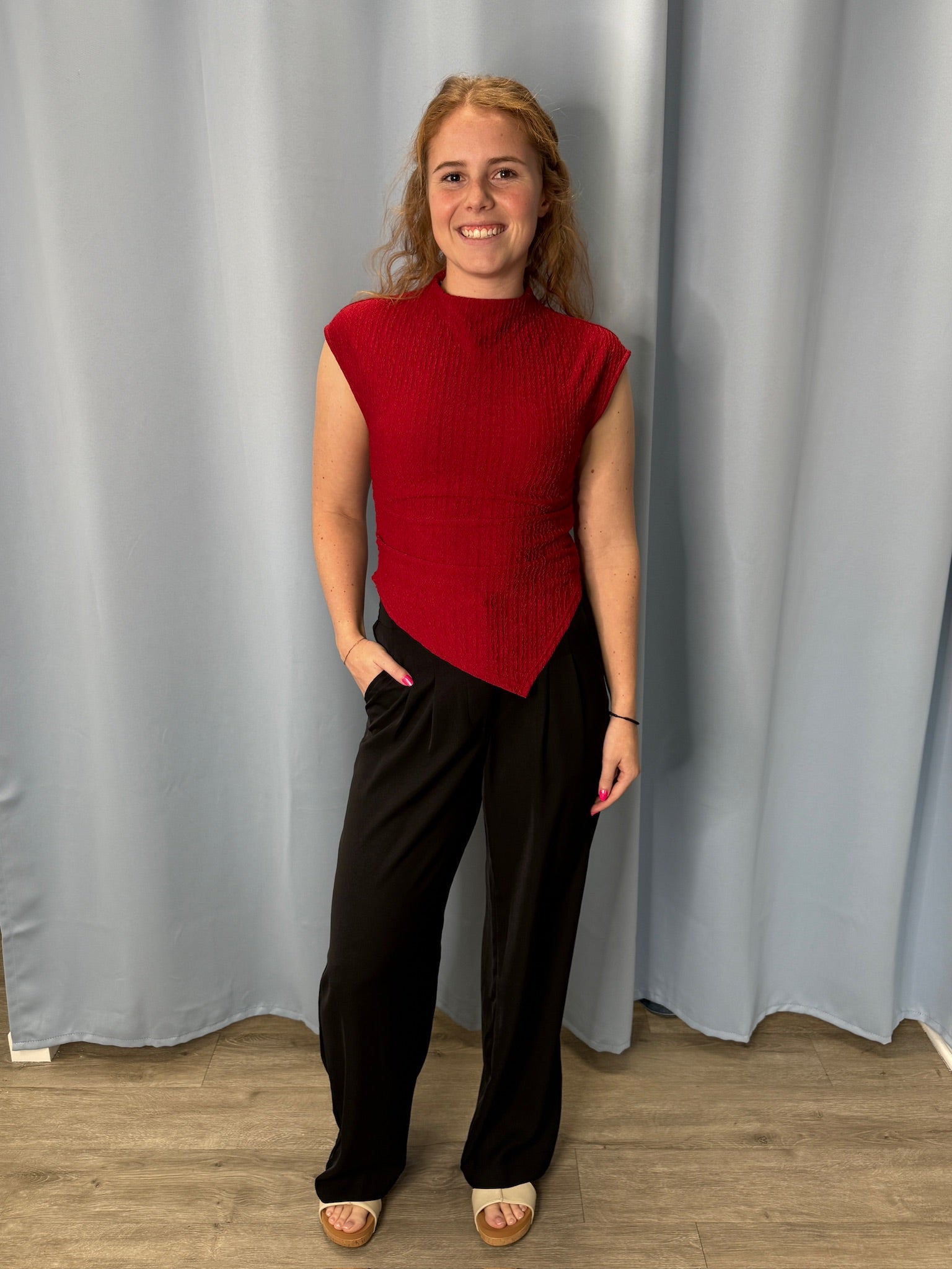 Person wearing a red top and black pants standing against a gray curtain.
