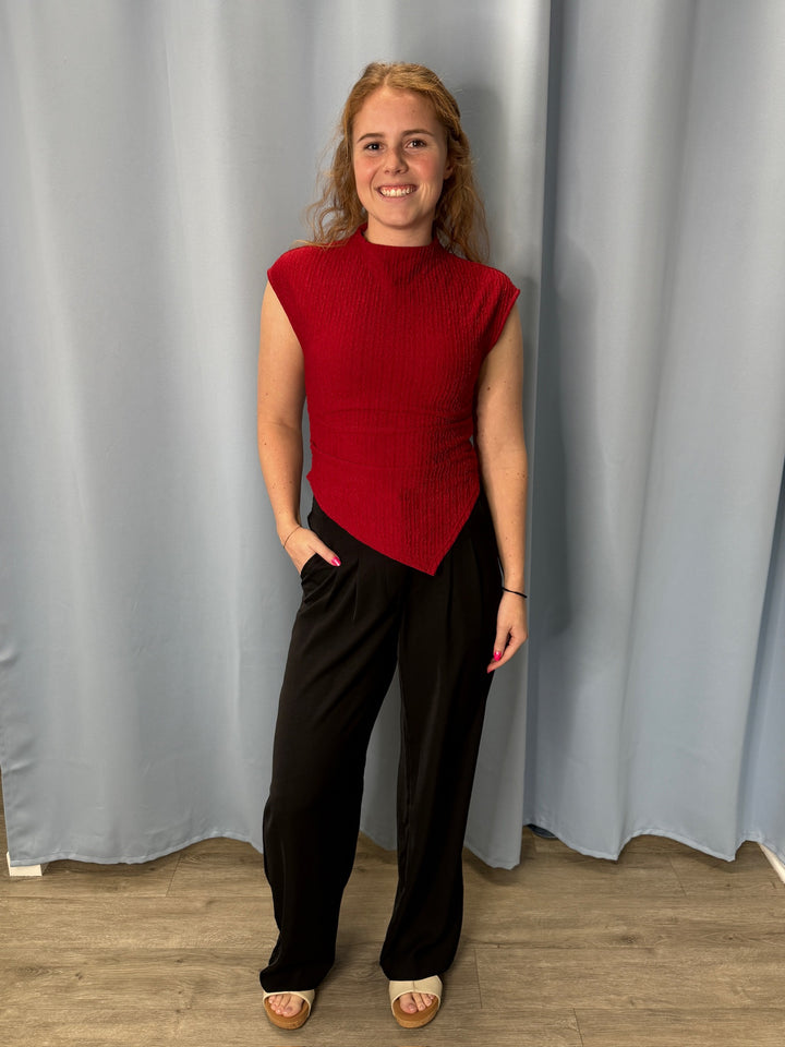 Person wearing a red top and black pants standing against a gray curtain.