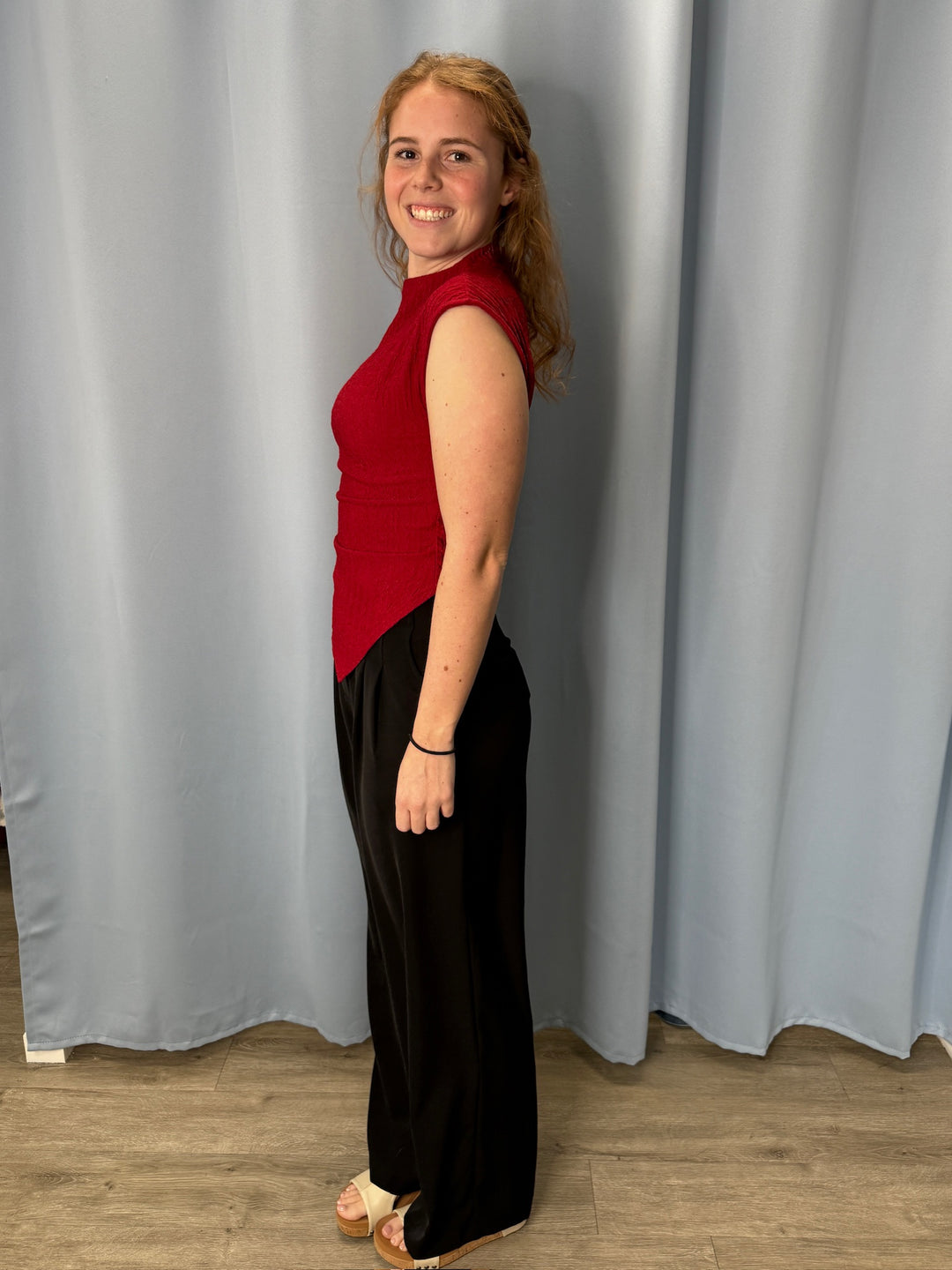 Woman wearing a red top and black pants standing against a gray curtain.