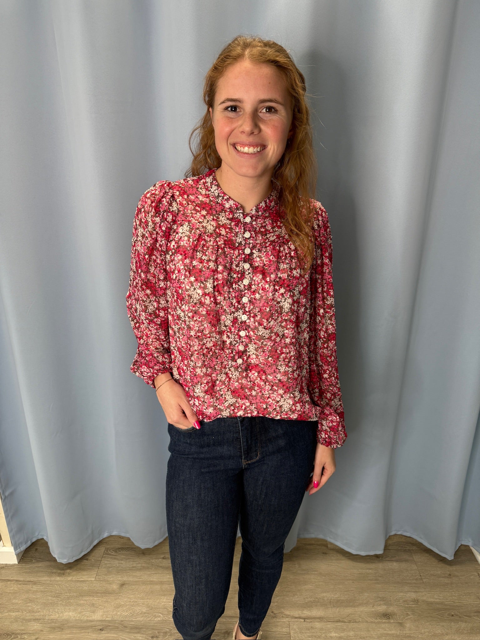 Woman wearing a pink floral blouse and dark jeans standing against a plain background