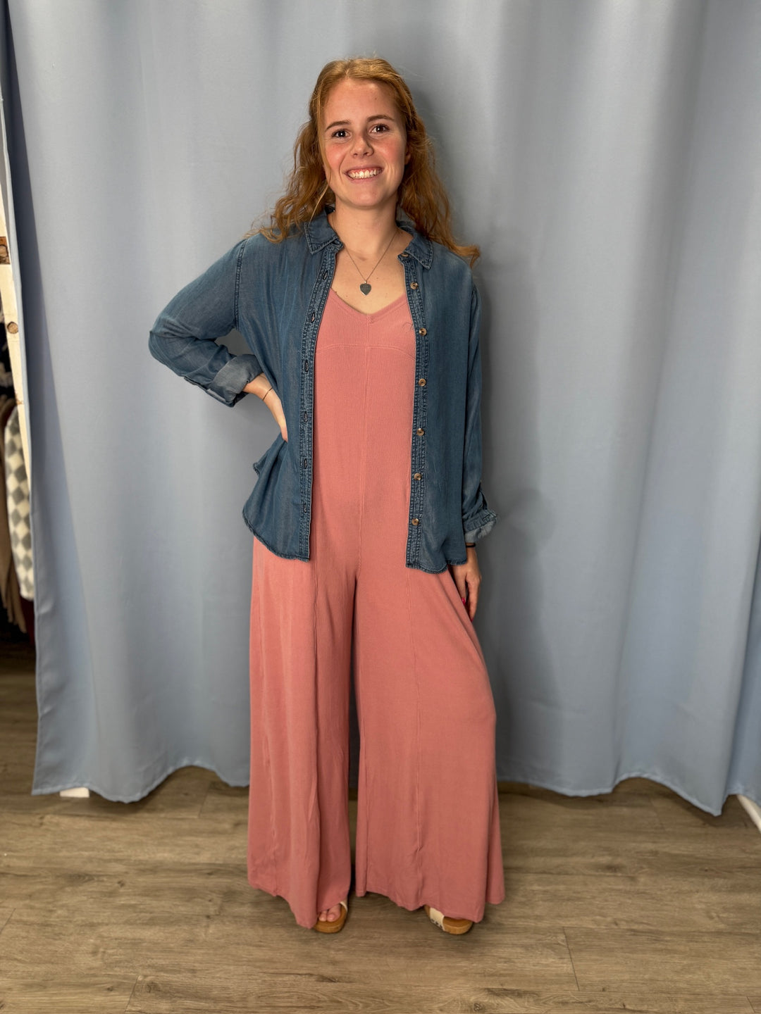 Woman wearing a pink jumpsuit and denim jacket standing against a plain curtain.