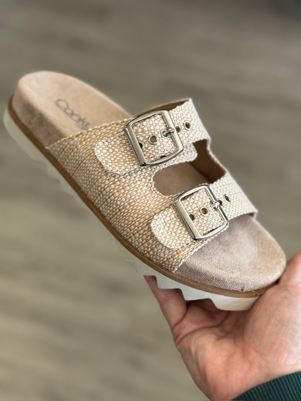 Beige sandal with two buckles held by a hand against a blurred background