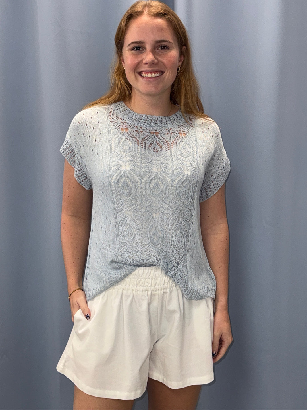 Woman wearing a light blue lace top and white shorts against a plain background