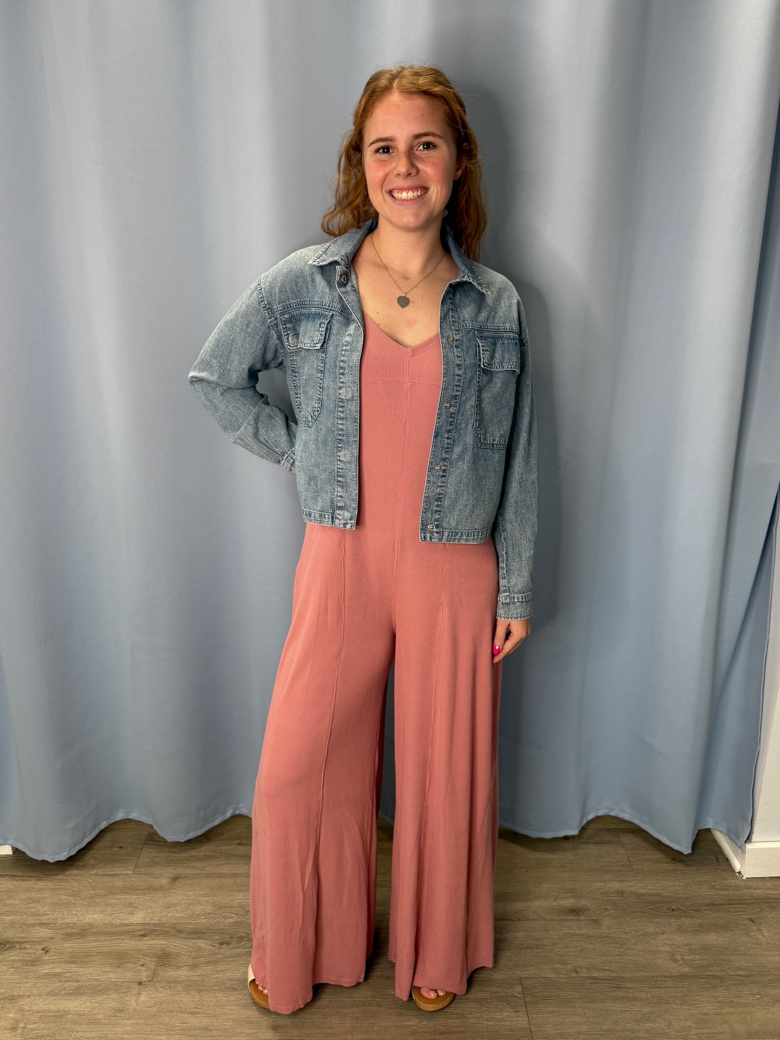 Woman wearing a pink jumpsuit and denim jacket against a gray curtain background