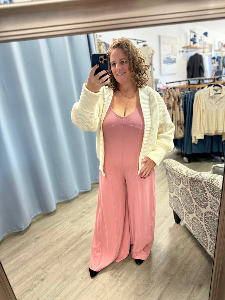 Woman taking a mirror selfie wearing a pink jumpsuit and white cardigan in a store.