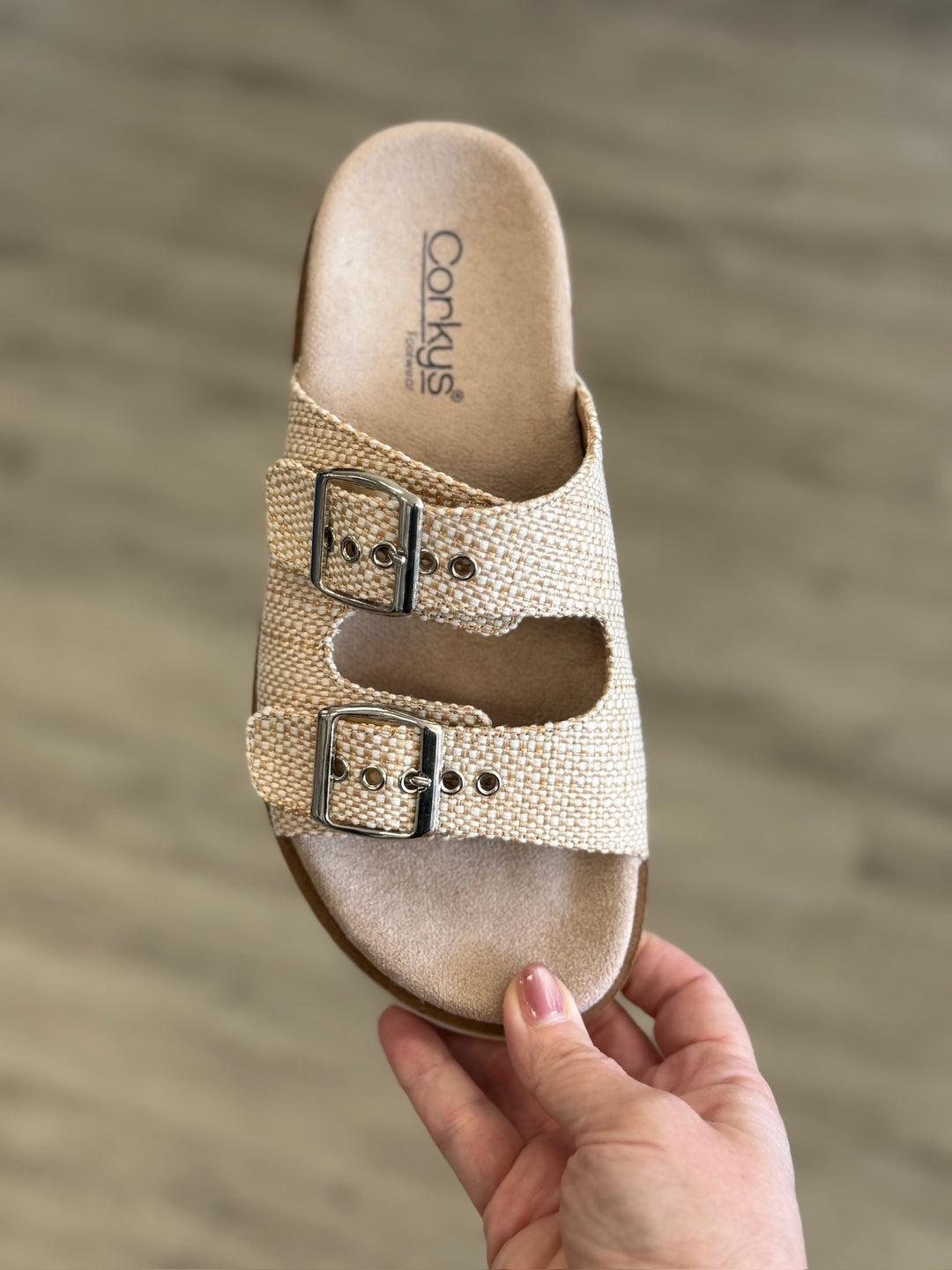 Beige sandal with two buckles held by a hand against a blurred background