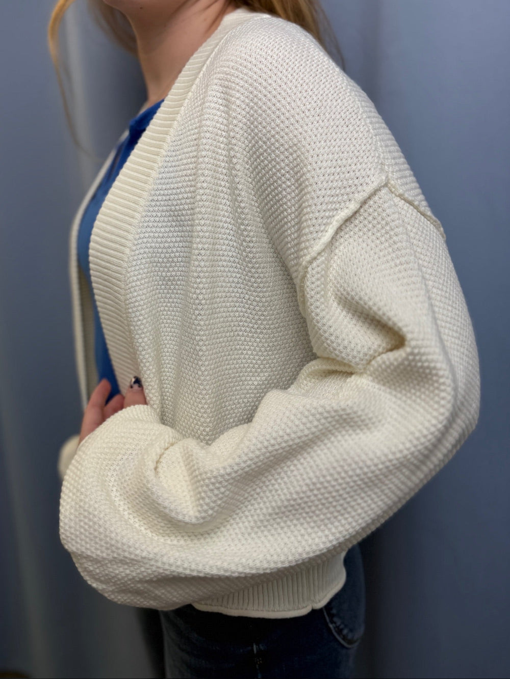 Person wearing a white textured cardigan over a blue shirt against a plain background