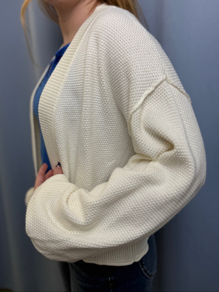 Person wearing a white textured cardigan over a blue shirt against a plain background