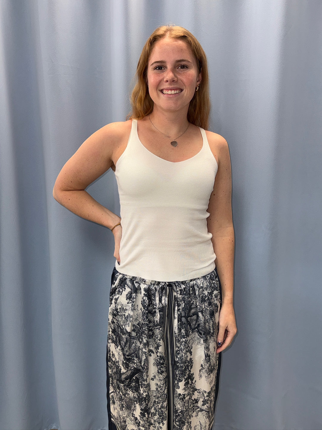 Woman wearing a white tank top and patterned pants standing against a plain background