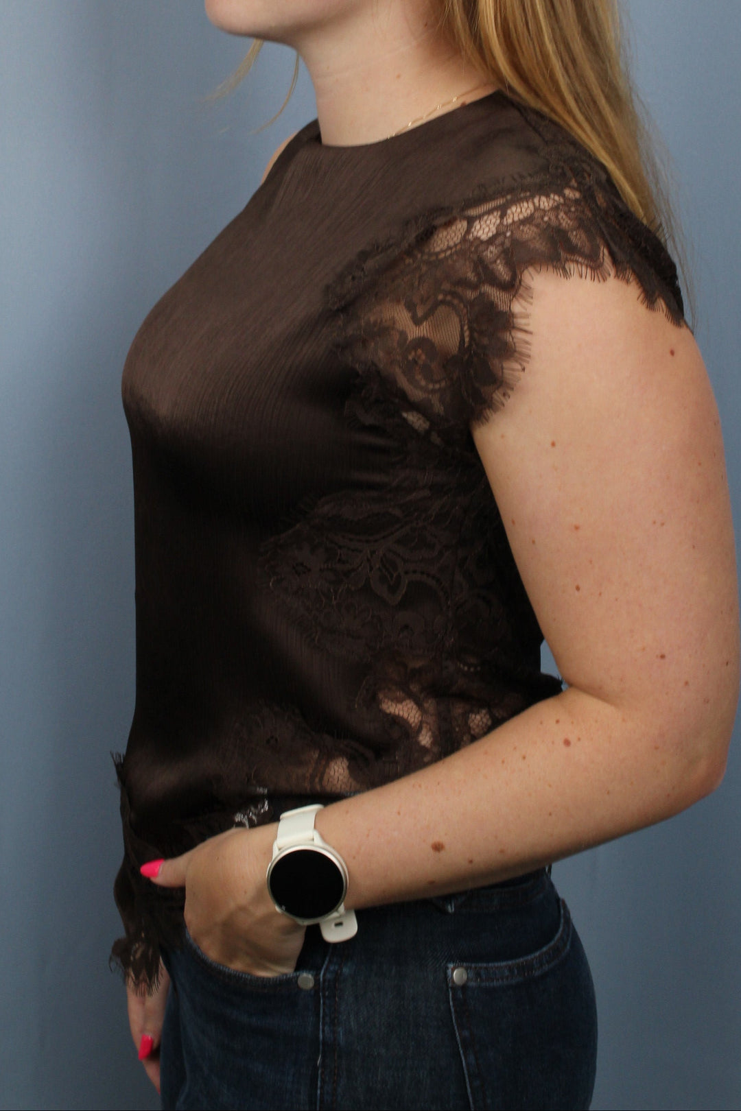 A woman wearing a sheer dark brown lace-trimmed top and dark jeans, posed against a solid blue-gray studio background.