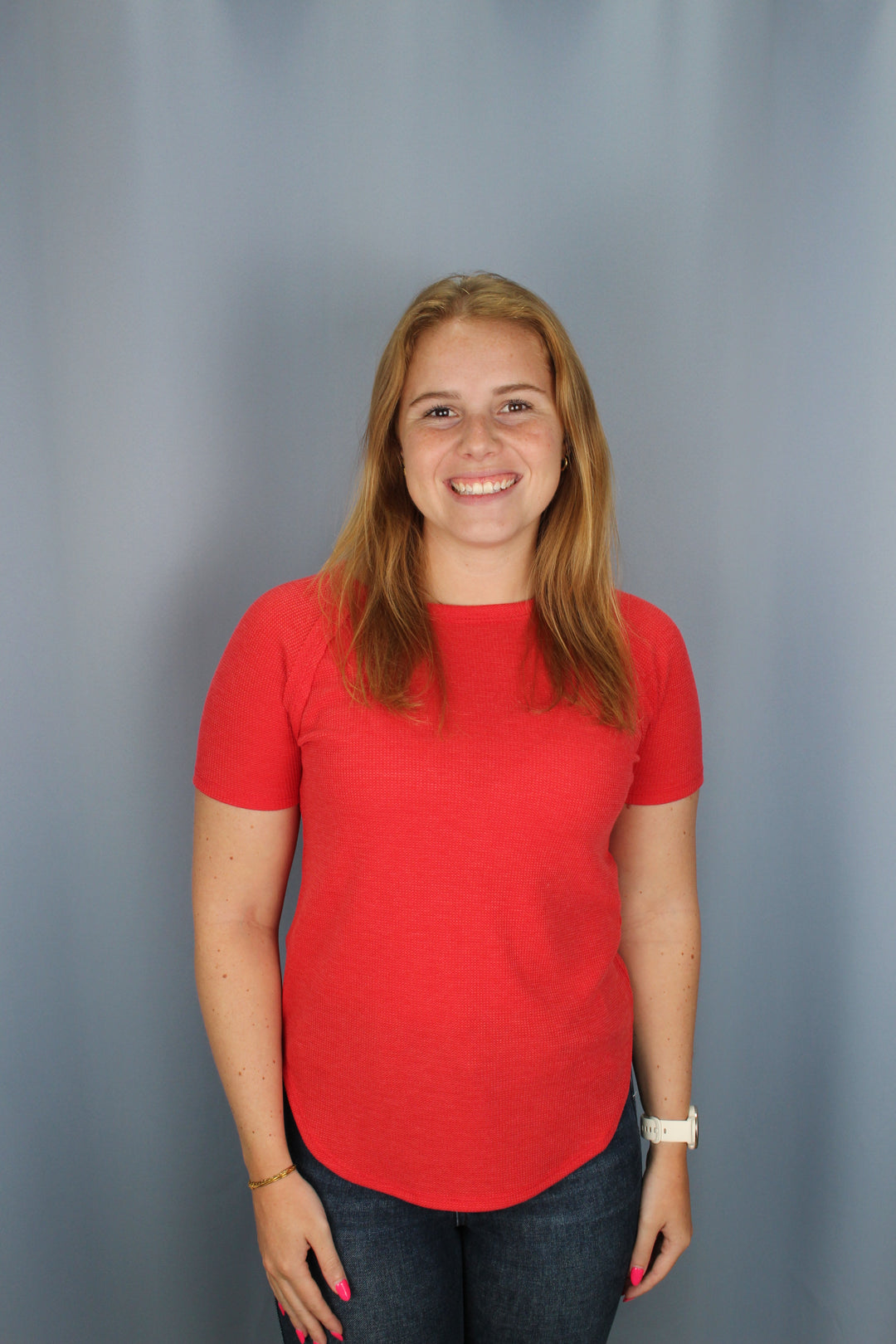A smiling young woman with long light brown hair is shown from the waist up, wearing a bright red short-sleeve t-shirt and dark blue jeans against a plain gray studio background.