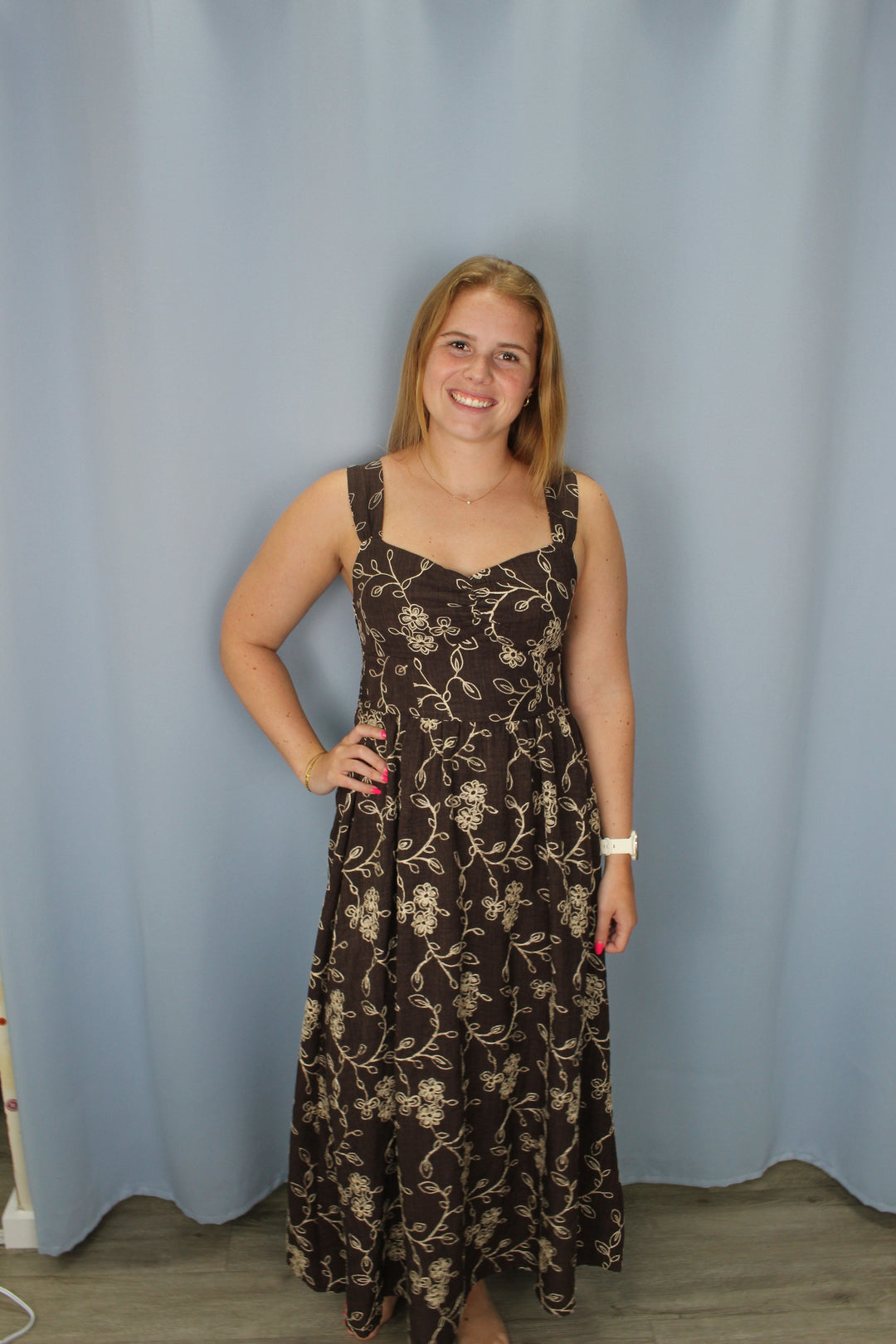 A woman wearing a brown sleeveless dress with a beige floral pattern, standing against a light blue backdrop.