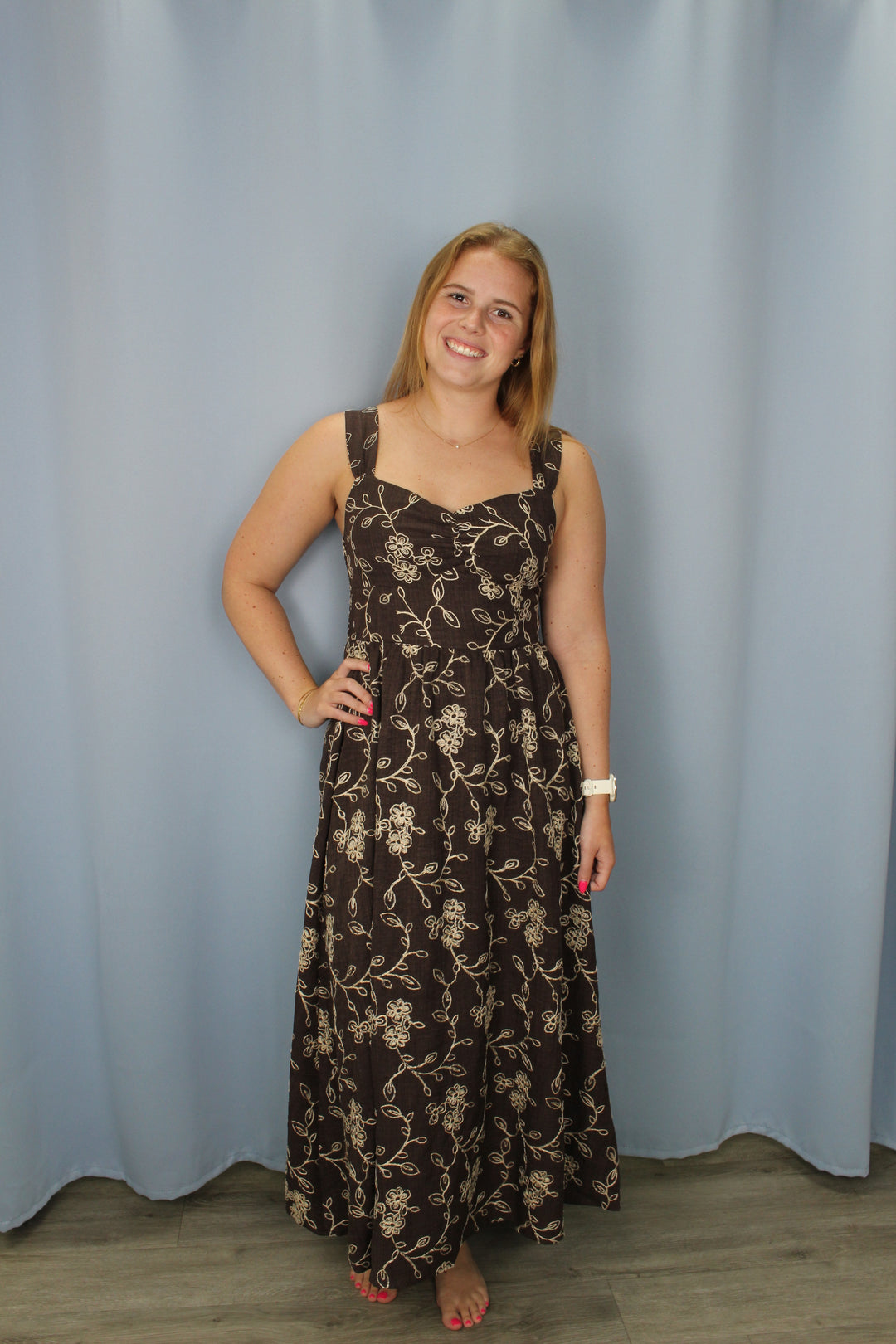 A woman wearing a sleeveless brown maxi dress with a beige floral pattern, standing against a light blue backdrop.
