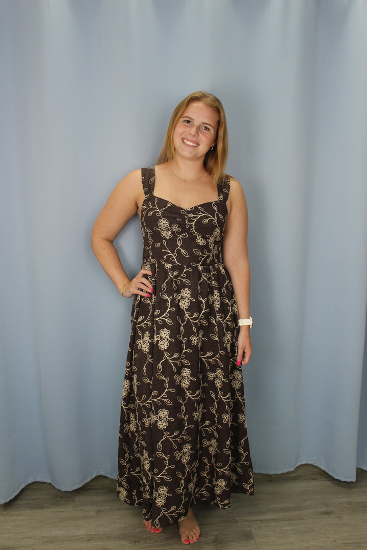 A woman wearing a sleeveless brown maxi dress with a beige floral pattern, standing against a light blue backdrop.