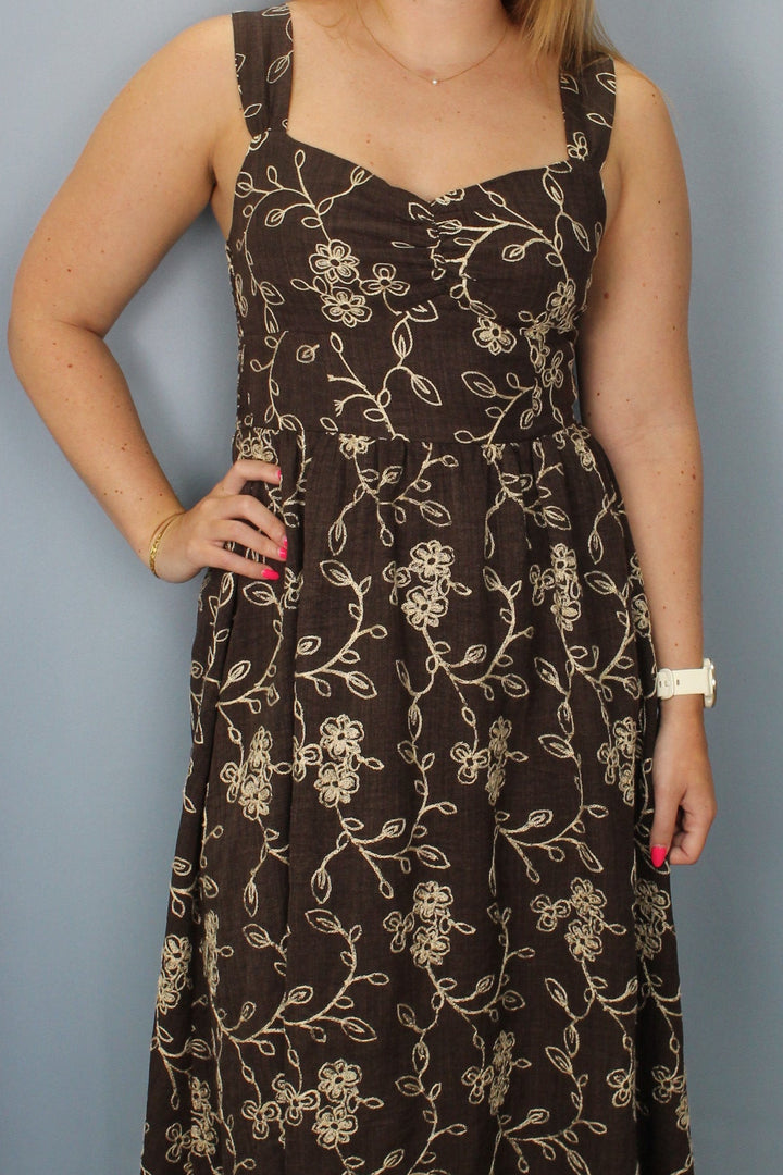 A woman modeling a brown sleeveless maxi dress with a beige floral vine pattern, standing against a light blue-gray backdrop.