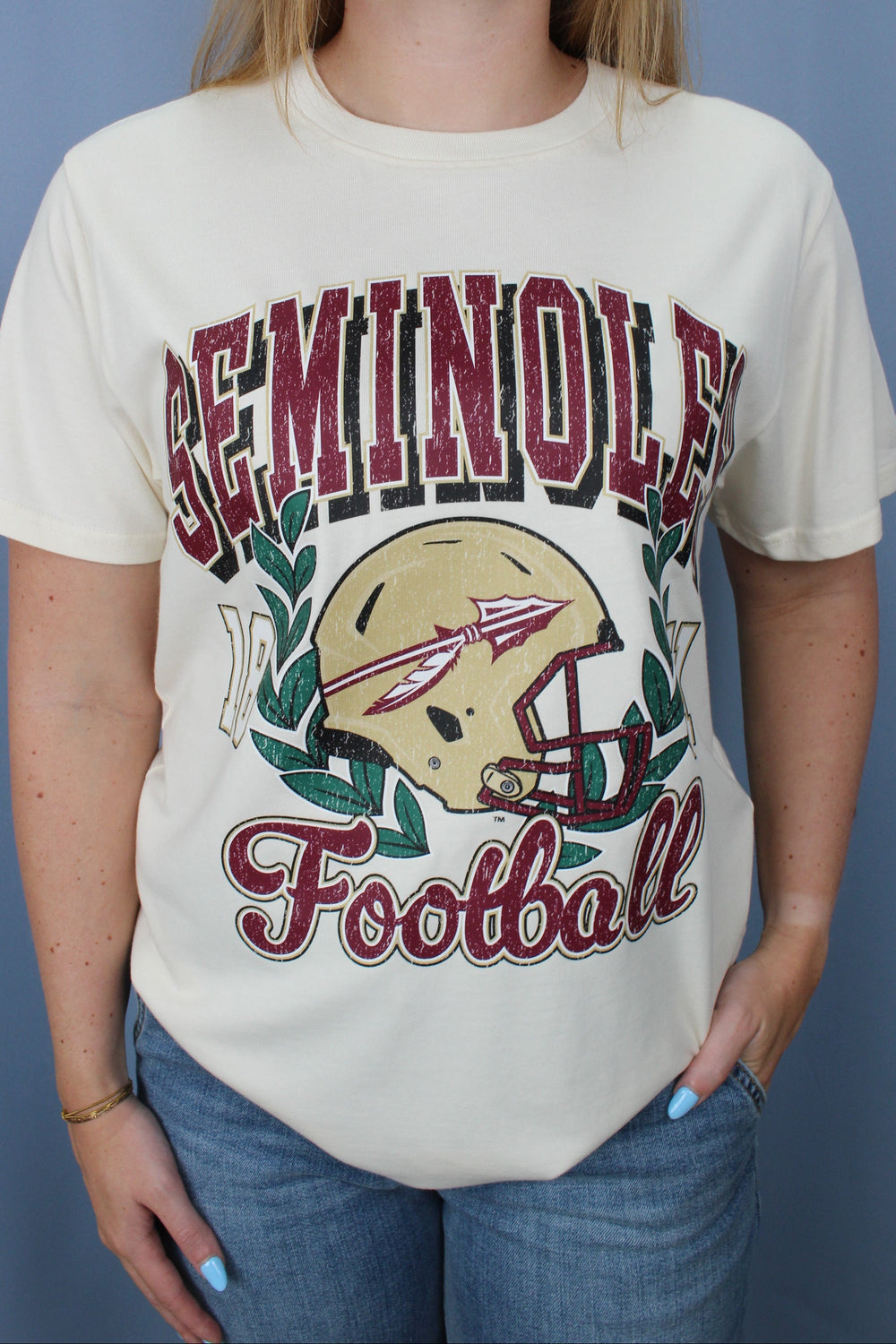 A woman wearing a vintage cream-colored Seminoles Football t-shirt with a graphic of a football helmet and the word 'SEMINOLE' in red and black, paired with blue jeans against a blue-gray background.