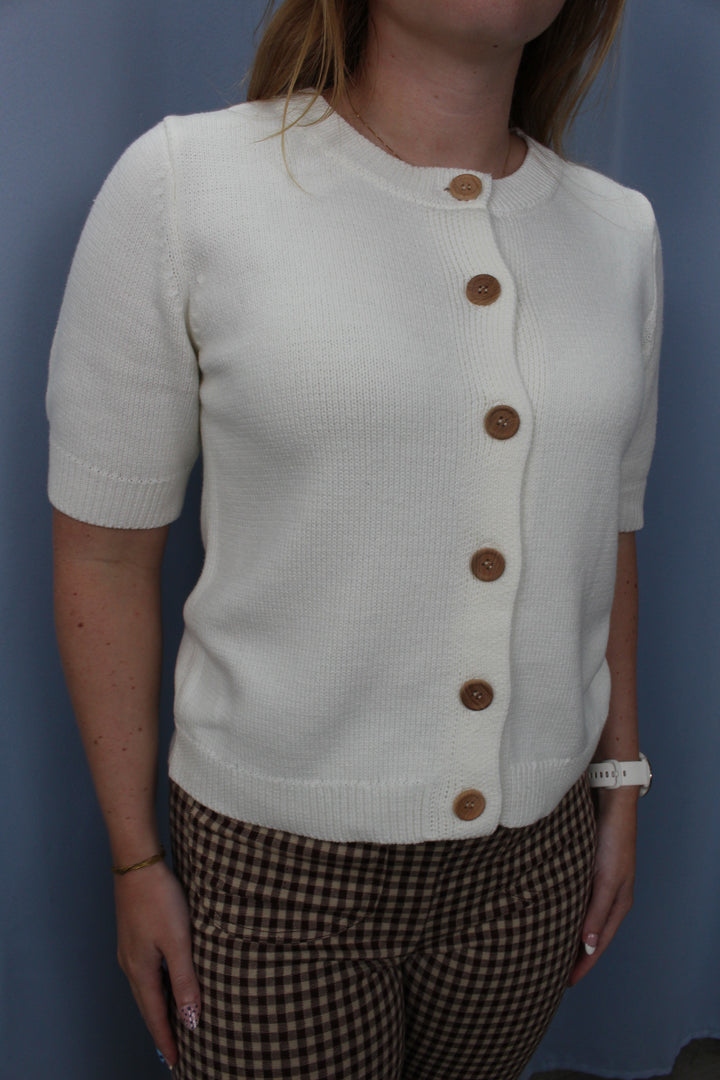 A woman wearing a short-sleeve cream knit cardigan with brown buttons, paired with brown and beige gingham pants, standing against a blue wall.