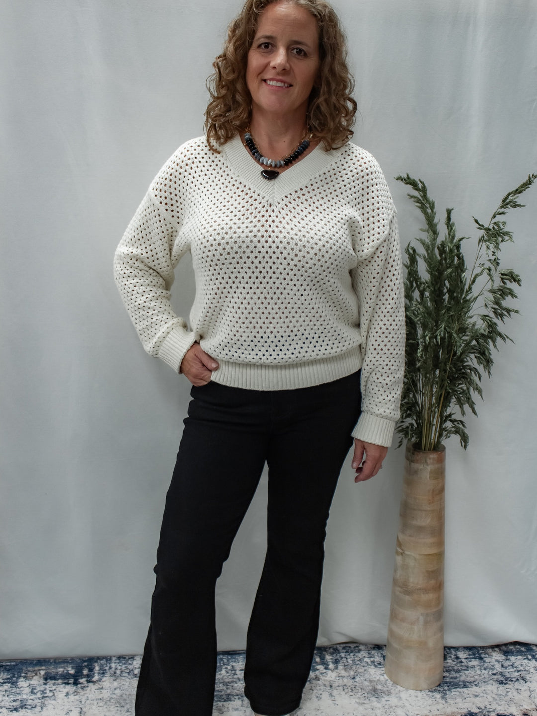A woman modeling a cream-colored, open-knit V-neck sweater with black pants, standing in front of a light gray backdrop next to a tall vase with greenery.