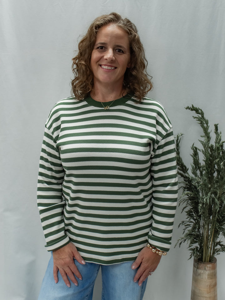 A smiling woman with curly brown hair stands against a light gray backdrop wearing a green and off-white horizontally striped long-sleeve top with light blue jeans. A potted dried plant is visible to her right.